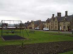 A village green with a swing set in the middle and a row of houses in the background