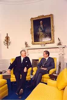 Whitlam sits in a chair, smiling, with Richard Nixon (whose appearance is well known) in another, who is also smiling.