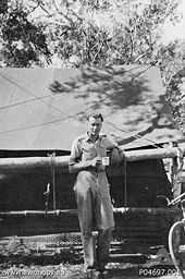 Whitlam in military uniform stands under a tree in front of a large tent.  He holds a mug in his hand.