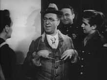 a man in what appears must be a brightly colored suit and hat, as this is a black and white photo, is the centre of attention speaking to three young men