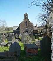 A simple church, seen end-on, with a bellcote and transepts; in the foreground are gravestones