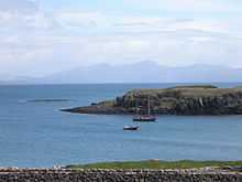 A two-masted yacht and a RIB lie at anchor on a sunny day off  a grassy shore, with low cliffs beyond. A skerry lies further offshore to the left with high hills in the distance.