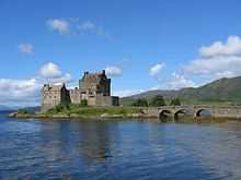 A stone castle in medieval Scottish style sits on a small island. An arched bridge leads off to the left.