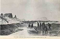 Nine men in dark clothing stand or sit around a small wooden boat that has been dragged on to a rocky shore, with rocks and icy peaks in the background.