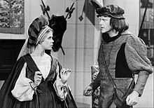 A black-and-white photograph of two people, a woman on the left and a man on the right, both standing and looking at each other while wearing poofy hats