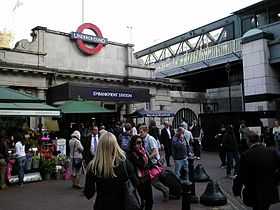 Single-storey white stone building with Underground roundel on façade above station entrance. Many pedestrians circulate in front of the station and a railway bridge fills the upper right portion of the frame