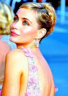 A young blonde woman with her hair tied up at the back, wearing diamond earrings and a pink flowery dress, photographed from behind and to the left and looking sideways from the viewer