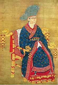 A painting of a woman in a blue dress with intricate gold and red decorations and a large blue hat. Her face has a simple angular design painted onto it in darker brown tones. She is sitting in a golden throne with dragon heads protruding from the ends of the armrests and from the sides of the top, back edge of the throne.