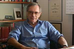 A grey-haired bespectacled man wearing a light blue shirt and sitting on a chair behind a desk