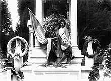 A woman in a long robe stands in front of a cemetery memorial, holding a large flag in her right hand, and raising her left arm.