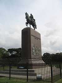 Behind a black iron fence stands a tall, rectangular stone plinth topped by a bronze equestrian statue