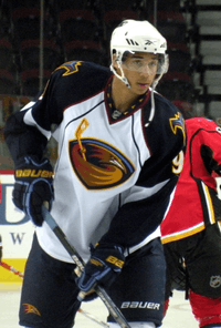 Hockey player in white and black uniform, with a picture of a bird's head in the middle. He leans forward slightly, holding his stick.