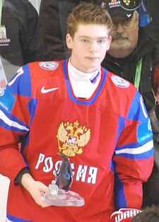 A short- and light brown-haired caucasian male in a red, Russian, ice hockey uniform is holding a small glass award.