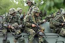  Six soldiers dressed in camouflage and face paint sit on top of a green vehicle.