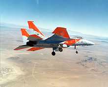 Jet aircraft with distinctive orange markings banking left over desert, with landing gears extended.