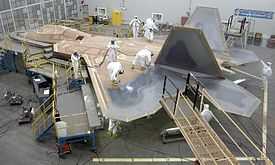F-22 being painted. Workers wearing white apparel standing on the aircraft's top applying a gray and black coat over the F-22. Temporary construction equipment surrounds its leading edges and nose sections.