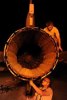 View of engine exhaust nozzle as two maintenance crew inspecting and repairing an F-16 engine at night
