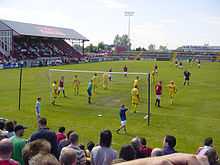 A football game in progress between F.C. United in red and Leigh Railway Mechanics Institute in yellow. The F.C. United crowd watch on.