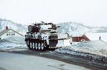 Vehicle in white winter camouflage. House roofs can just be seen behind and in the distance a range of snow covered mountains