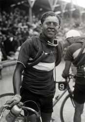 A man with a dirty face holding his bicycle, spare tires wrapped around his shoulders. In the background a large crowd.