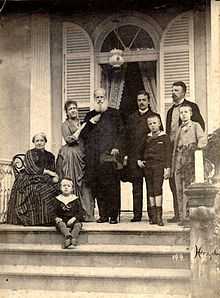 Photograph of a group of people assembled on a columned porch at the top of a flight of steps, with one older lady seated, one younger lady leaning on the arm of an older bearded man, two younger men and three small boys