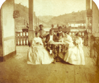 a man and three women seated around a table with two servants standing in the background