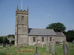 Gray stone building with square tower at left hand end. Grass and gravestones in the foreground.