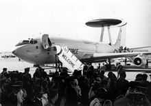 Black-and-white photograph with angled front view of four-engine jet aircraft on ramp with front fuselage door opened. A contingent of people are there to welcome the jet, which has a disc-shaped radar perching on top of struts on the dorsal fuselage.