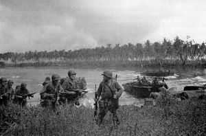 Soldiers walk through long grass. Other soldiers are arriving in landing craft in the lagoon behind them. In the background is a coconut plantation. The sky is overcast.