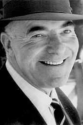 A black-and-white photograph of a man looking slightly to the right of the viewer and smiling while wearing a hat and a necktie