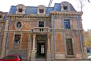 A three-story brick building with a balcony supported by stone pillars over the main entrance and stone trim generally. The upper story is a mansard roof pierced by dormer windows with round-arched roofs. Some of its windows are shattered and it appears abandoned, but a small maroon car is parked at the lower left and utility wires enter the building through the central window on the upper story