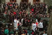 A crowd of mourners standing at the steps leading to the entrance of a church, facing the street. A hearse is partially visible at the bottom right of the photograph; beside it is a casket, being carried to the hearse by eight pall bearers, and being trailed by the family of the decedent.