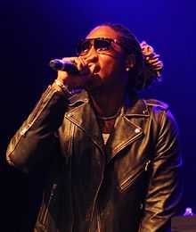 A portrait of an African-American man with dreadlocks, performing on stage, wearing a jacket and sunglasses.