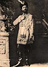 Monochrome photograph of a man standing, wearing leather books, knee-length tunic and feathered hat
