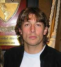 A photograph of a man with medium-length brown hair wearing a black jacket over a white shirt.