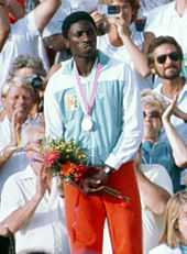 An African athlete wearing a light blue and white top and red shorts. He has a silver medal hanging from his neck and holds a posy of flowers.