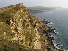 A large, rocky cliff overlooking the sea to the right, crashing into the shore.
