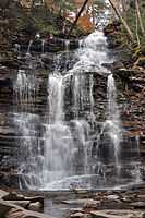 Head-on view of a wide and highly vertical waterfall from near its base. Large, jagged boulders protrude into the foreground and continue toward the falls. Two hikers standing on a ledge near the top are dwarfed by the scope of the falls.