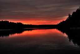 Red, yellow, and purple clouds are reflected in a lake surrounded by silhouetted trees.