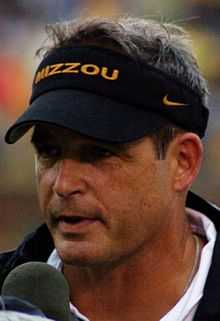 A headshot of a man with a microphone to his mouth wearing a black visor with the word "MIZZOU" in gold on the front.