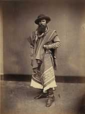 Full-length photograph of a bearded man wearing a wide-brimmed hat, bandana, serape, leather boots and grasping the handle of a long knife in his left hand