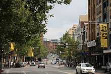 A photograph of a street with motor vehicles travelling in both directions and buildings on either side all under a blue sky with white clouds