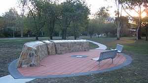 An ellipse of orange pavers surrounded by a grey boarder, with a man-made rock structure on one side of the ellipse displaying plaques. On the other side are two bench seats. Grass surrounds the ellipse and trees can be seen in the background.