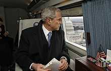 George W. Bush wearing a suit, tie, scarf and overcoat while looking out the window from a skyward vehicle.
