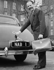 Full length picture of a middle aged man with a very large, bushy mustache. He is wearing a suit and carrying a briefcase. He is standing at the rear of a parked car with his hand on the boot-handle.