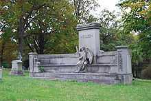 Short, wide memorial lined by a bench with angel statue sitting on the bench. Material is grey and central pillar depicts the name Giles.