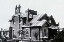 Black and white photograph of the facade of the brick house. Two stories with a two-pronged chimney rising up the front wall.