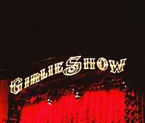 Image of a concert stage. It is written "GIRLIE SHOW", in yellow, above red curtains, as flashes of lights illuminate the stage.