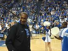 A man, wearing a black jacket and black t-shirt, is walking onto the basketball court.