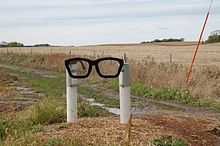 A sculpture of sort, consisting of two white posts holding a black spectacles frame in Buddy Holly's characteristic style